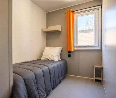 Chambre simple avec lit, petite tablette et rideau orange dans le mobil-home Le Hameau des Pins