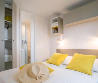 Chambre avec lit, oreillers jaunes et chapeau de paille au Camping Le Marqueval
