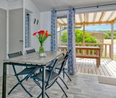 Salle à manger ouverte sur terrasse en bois avec table et vue jardin du Camping Le Village de la Mer