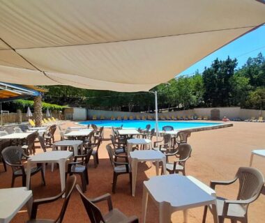 Terrasse ombragée avec tables et piscine en arrière plan au Camping Naturiste Tikayan Petit Arlane