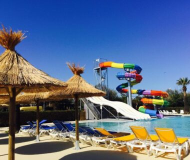 Piscine avec toboggans colorés, transats et parasols en paille au Camping Resort Les Champs Blancs