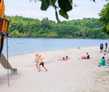 Plage du lac avec aire de jeux et baigneurs au Camping du Lac