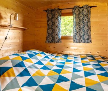 Intérieur d'une cabane en bois avec lit couverture géométrique et petite fenêtre au Domaine de la Cambre