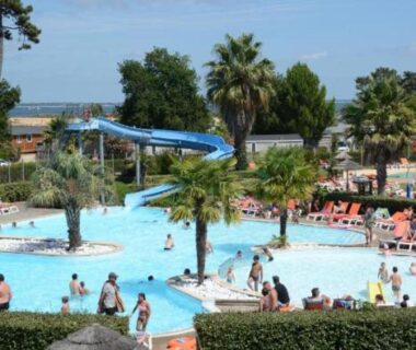 Piscine extérieure avec toboggan et palmiers au camping ESCALE BASSIN Village 4 étoiles LES VIVIERS