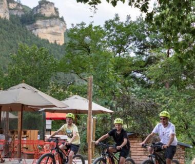 Sous les arbres trois cyclistes devant une terrasse au camping Huttopia Gorges du Tarn