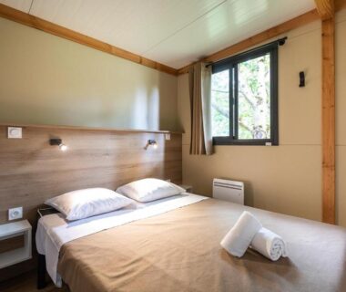 Chambre avec lit double serviettes pliées et fenêtre dans le lodge Huttopia Sarlat