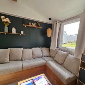 Séjour avec canapé d'angle et étagère décorative près de la fenêtre du Jolie Mobil home 2 chambres (198)