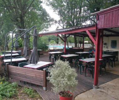 Terrasse couverte avec tables et chaises près de la piscine au camping L escale de larcher