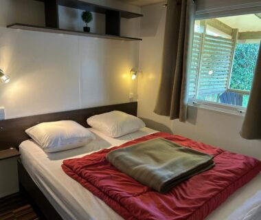 Chambre avec lit double lampes murales et fenêtre sur terrasse au camping LES PIERRES CHAUDES