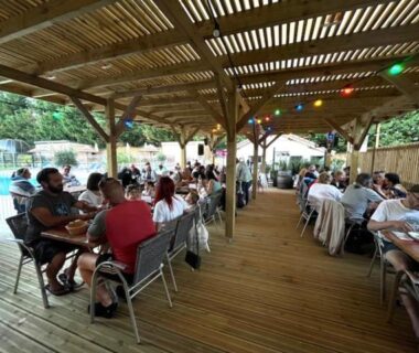 Terrasse couverte avec tables, convives et piscine en arrière-plan au camping L'imprévu