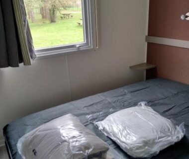 Chambre avec lit, oreillers emballés et fenêtre du Mobil Home