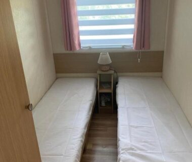 Chambre avec deux lits simples, lampe, rideaux et fenêtre à store dans le Mobil Home