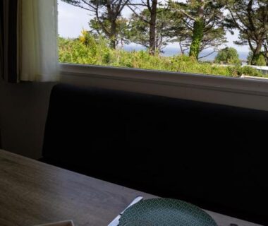 Table et viennoiseries devant fenêtre avec pins et vue sur mer du Mobil-home convivial avec vue sur mer Pleubian