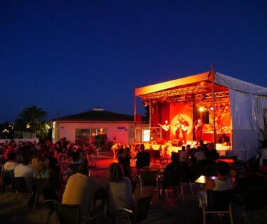 Scène extérieure illuminée et public assis en soirée dans le camping les sables du midi valras plage